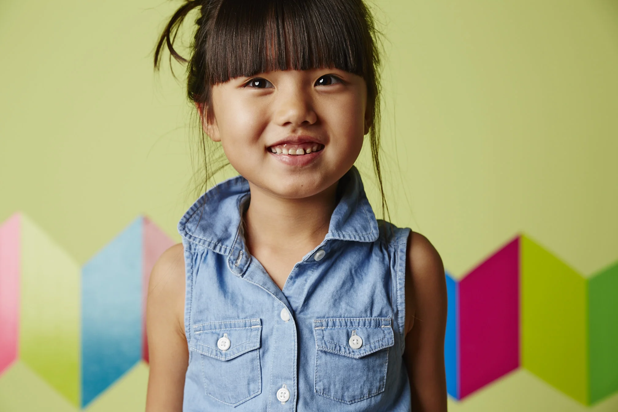 A young girl smiling with missing teeth, wearing a sleeveless denim shirt, standing in front of a colorful geometric background.