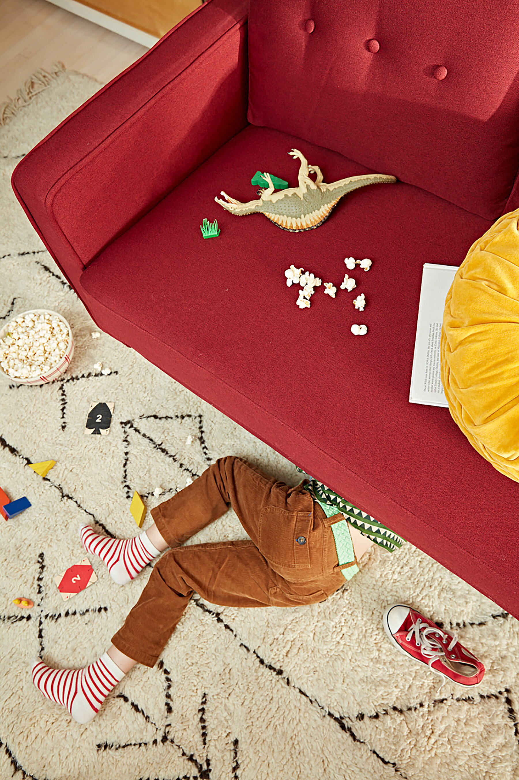 A child's legs are visible under a red couch, with one red sneaker and the other off. The child is wearing brown pants, a green and white patterned shirt, and red and white striped socks. On the carpet, there are scattered popcorn, a black number car