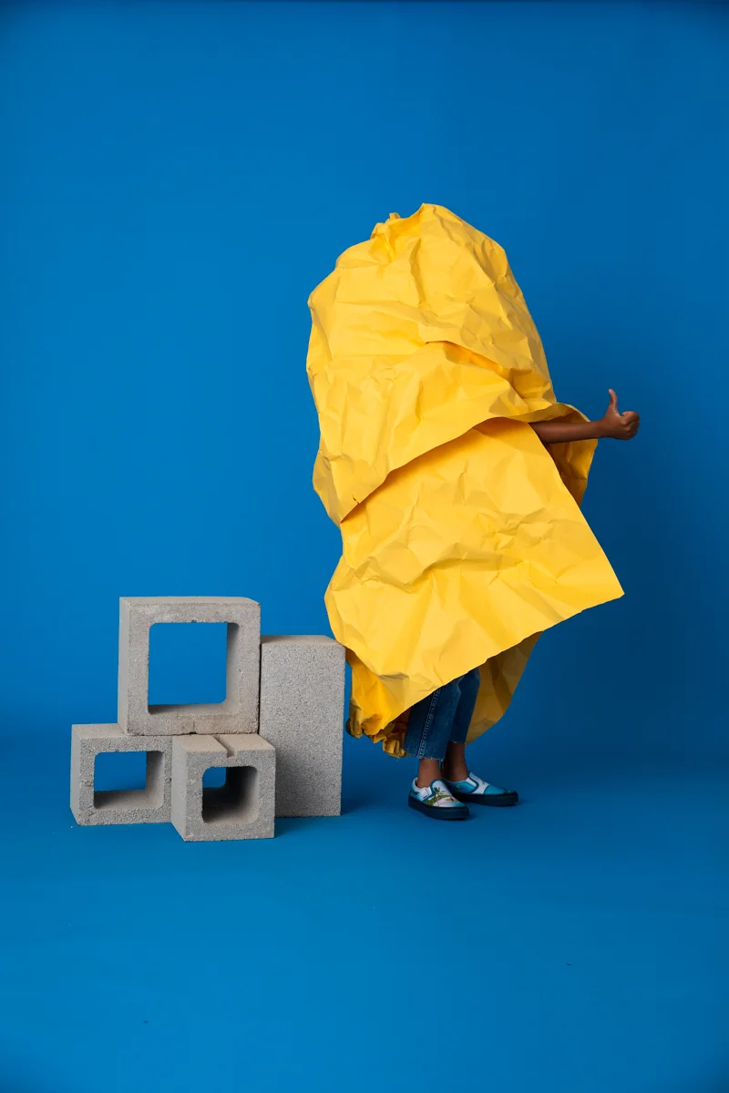 A person standing behind a large yellow paper umbrella, holding an arm out, with concrete blocks placed on the floor next to them, against a blue background.
