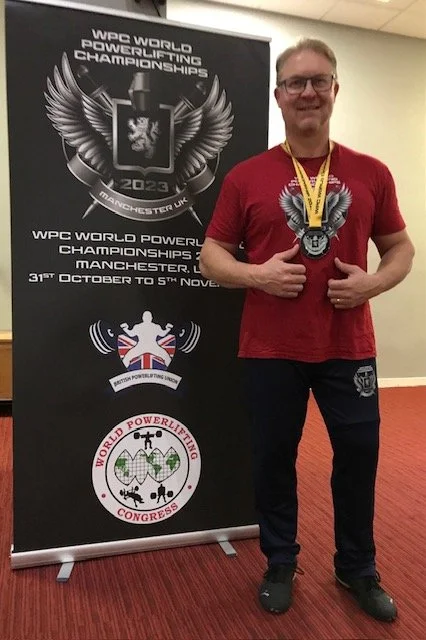 Man wearing a red T-shirt with a winged lion emblem, standing next to a black banner for the WPC World Powerlifting Championships in Manchester UK. He has a medal around his neck and is giving a thumbs-up.