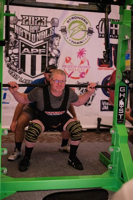 A woman in black shorts and a gray t-shirt is doing a squat with a barbell on her shoulders, assisted by a spotter standing behind her in a gym. The background has banners and logos related to powerlifting and strength training.