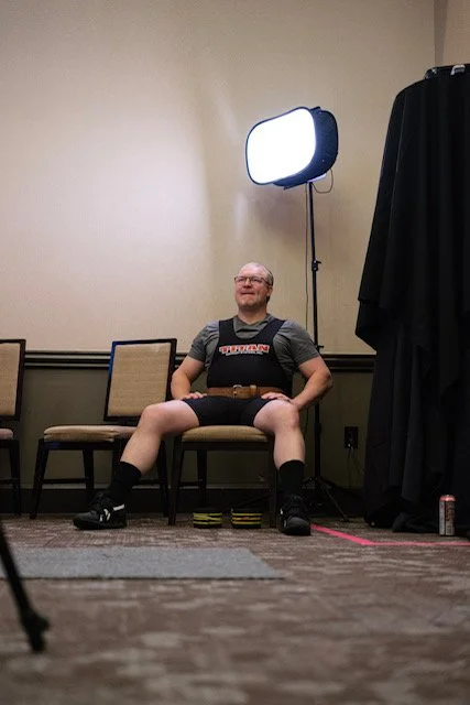 A man sitting on a chair with his legs spread apart, wearing dark shorts, a T-shirt, and a harness, smiling. There is a bright light on a stand behind him, a black curtain to the right, and chairs and a wall in the background.