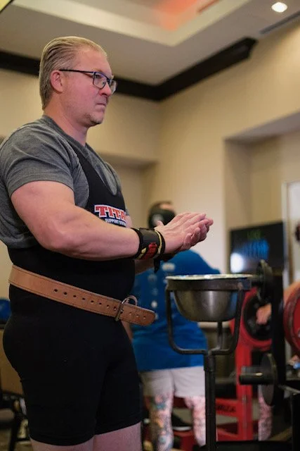 A muscular man with blonde hair and glasses, wearing a gray T-shirt with a logo, black shorts, a brown belt, and wristbands, is standing indoors near a weightlifting or fitness area.