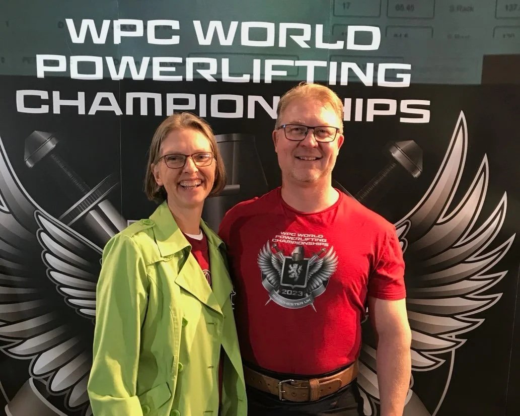 Two people standing in front of a backdrop that says 'WPC WORLD POWERLIFTING CHAMPIONSHIPS'. The woman on the left is wearing glasses and a bright green jacket, and the man on the right is wearing glasses and a red T-shirt with the event logo. They are smiling and posing for the photo.