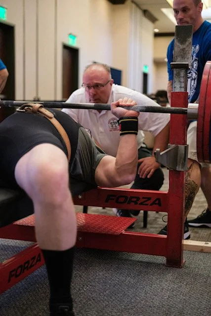 Man performing a bench press on a Forza bench press station, with two spotters assisting and observing in an indoor setting.