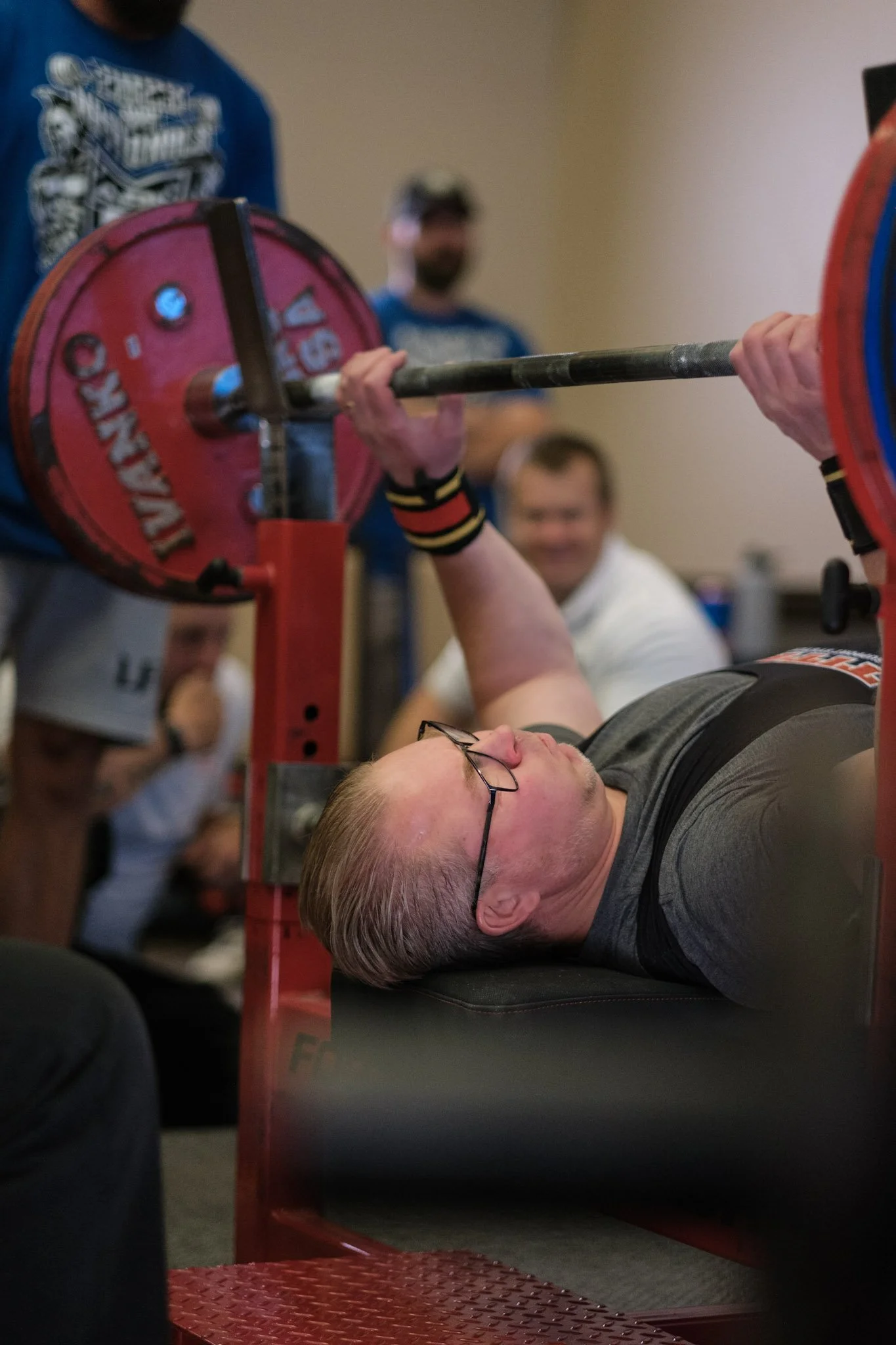 A man with glasses lying on his back on a flat surface, lifting a barbell with red weight plates during a weightlifting competition or training session. People are watching in the background.