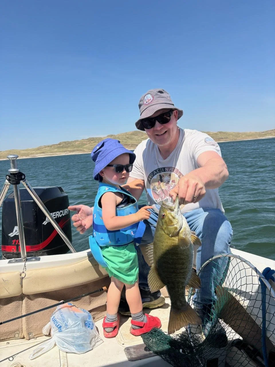 Man and young boy on a boat holding a large fish, with a body of water and distant landscape in the background. The man is smiling and wearing sunglasses and a hat, while the boy is wearing sunglasses, a blue hat, and a life jacket.