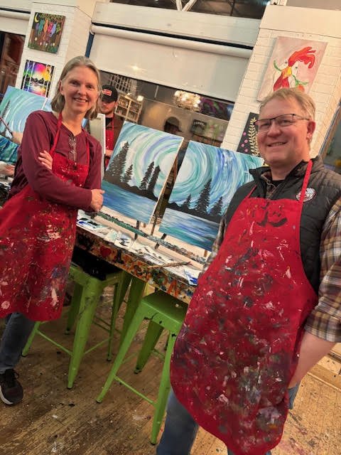 Nick and Annette wearing red aprons paint landscape canvases with northern lights over trees at an art studio, with art supplies and paintings in the background.