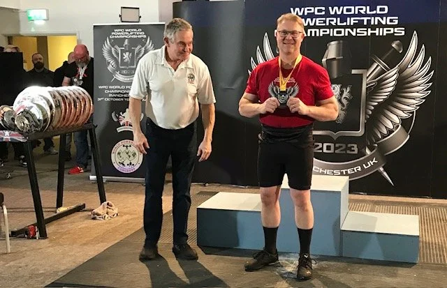 A powerlifter wearing a red shirt, black shorts, and black knee-high socks standing on a podium with a medal around his neck, holding a small trophy, smiling. An older man in a white shirt and black pants stands beside him, looking at the powerlifter