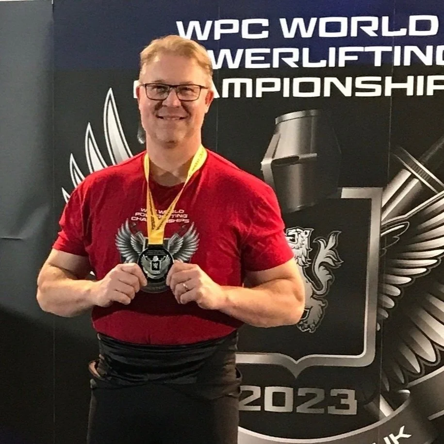 A man with glasses in a red shirt holding a medal at the WPC World Powerlifting Championship 2023.
