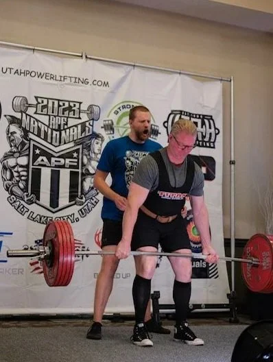 A man with glasses is performing a deadlift with a barbell loaded with large red weight plates at a powerlifting event. A spotter stands behind him, ready to assist. The background includes a banner with logos and text related to powerlifting and fitness.