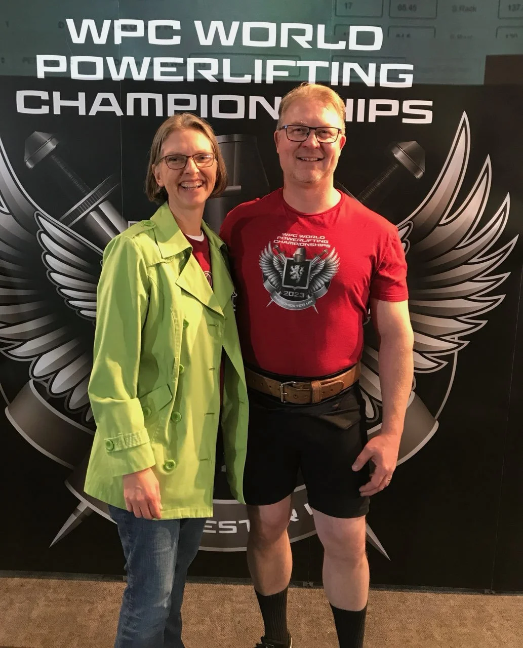 Smiling woman and man standing in front of a backdrop with the text 'WPC World Powerlifting Championships.' The woman is wearing glasses, a lime green jacket, and jeans. The man is dressed in a red T-shirt with a logo, black shorts, and black knee-hi