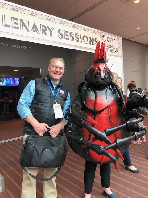 A man in a blue shirt and black vest standing next to a person in a detailed lobster costume at a conference. There is a banner overhead reading 'Lenary Sessions' and event details. The setting appears to be a conference hall.