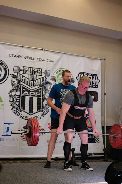 A man in black shorts and a gray shirt with weightlifting straps is performing a deadlift in a competition, while a spotter in a blue shirt assists and encourages him.