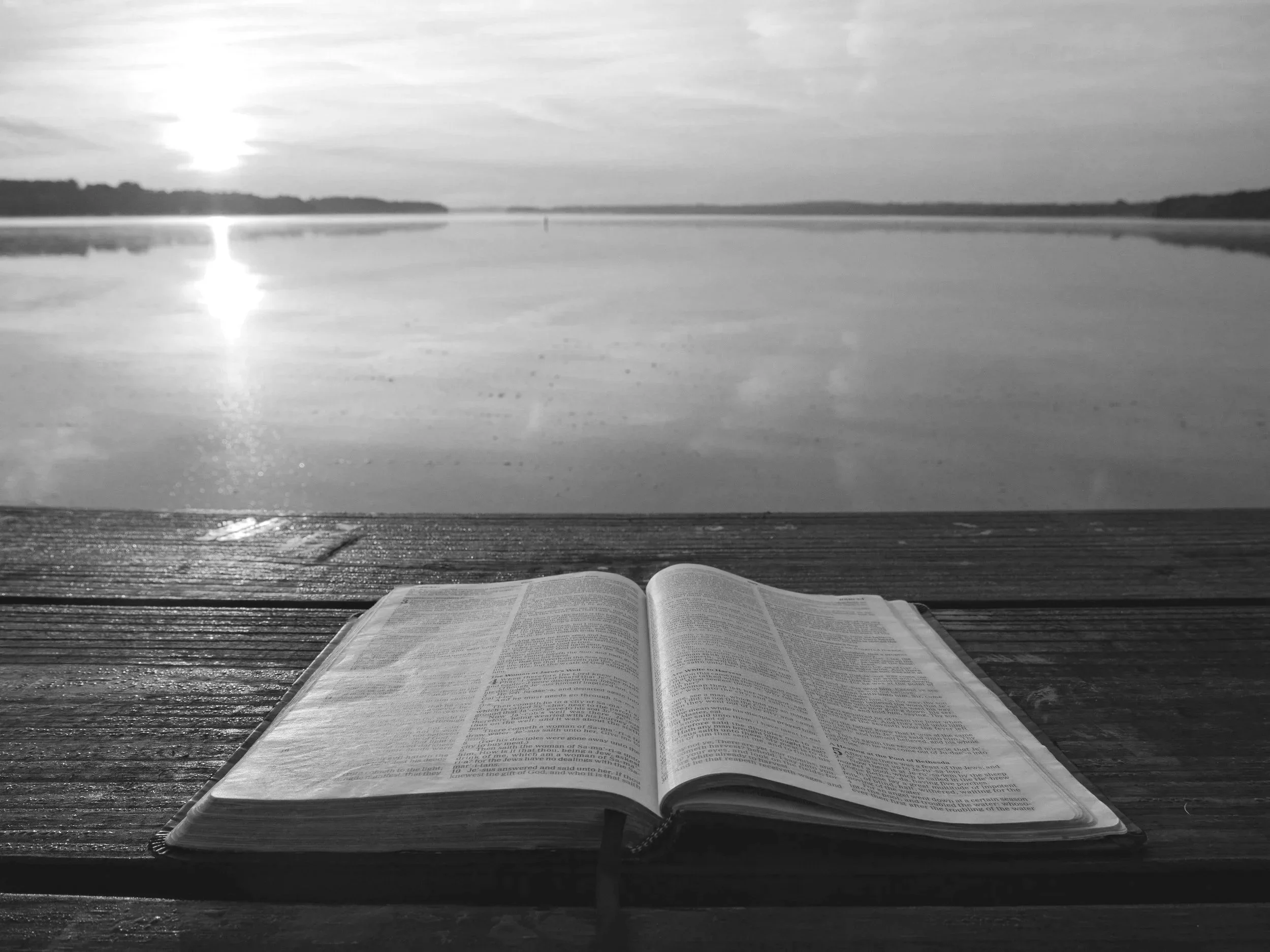 Open book resting on a wooden surface with a lakeside view at sunset or sunrise, calm water reflecting the sky, and trees in the background.
