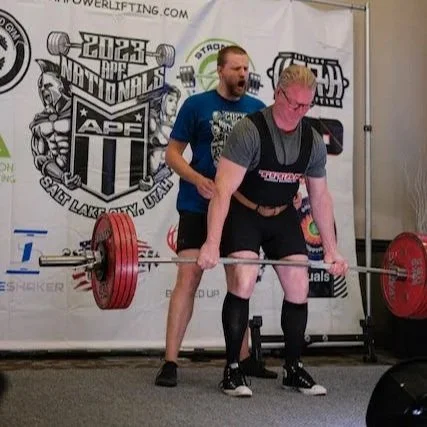 Nick performing a deadlift at a powerlifting competition. The backdrop features banners for the 2023 APF Nationals in Salt Lake City, Utah.