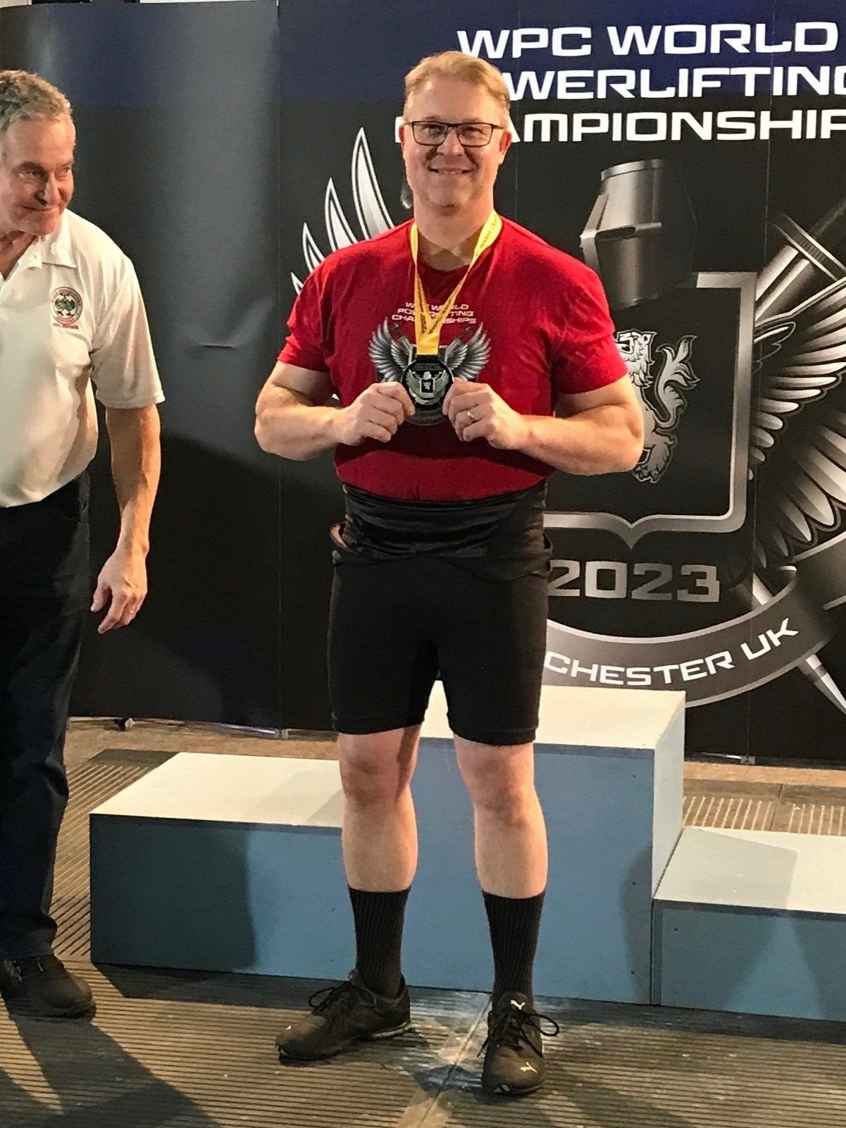A man with glasses, wearing a red shirt and black shorts, holding a medal, standing on a winners' podium at the WPC World Powerlifting Championship 2023 in Chester, UK, with another person partially visible on the left, and a dark backdrop with event