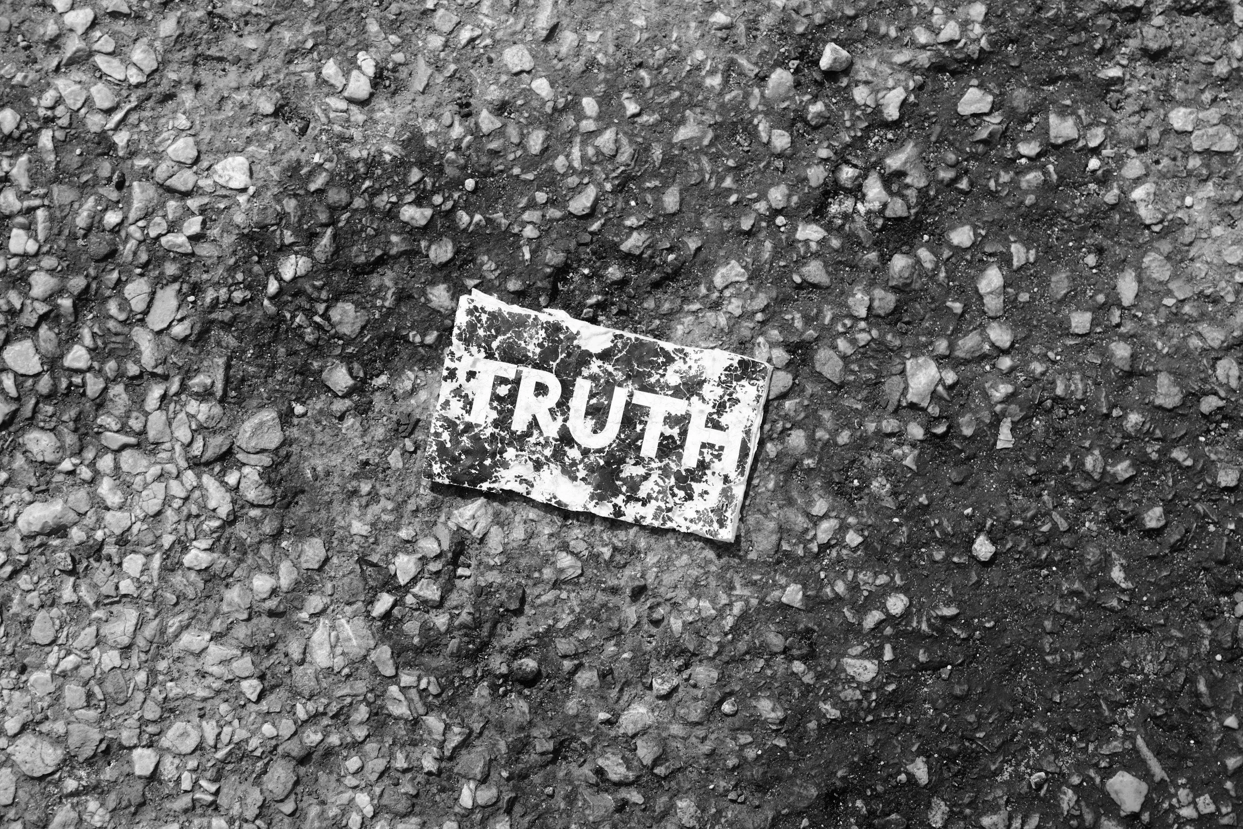 A close-up black and white photograph of gravel or small stones with a crumpled piece of paper on the ground that has the word 'TRUTH' printed on it.