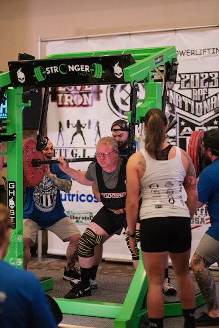 Man performing squat with weighted barbell inside a green powerlifting cage during a competitive event, surrounded by spotters and spectators.