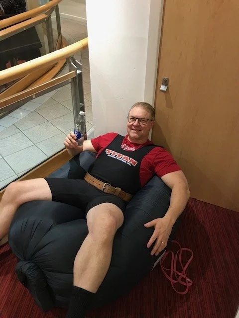A man lying on a black inflatable chair in a hallway, holding a blue bottle, smiling at the camera, wearing glasses, a red and black sports shirt, black shorts, and black socks.