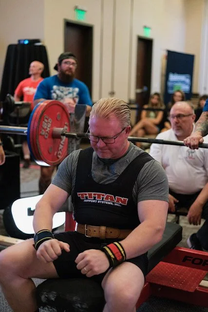 Man with glasses and blonde hair participating in a powerlifting competition, sitting on a bench with a barbell behind him.