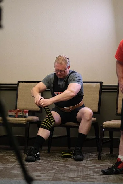 A man sitting on a chair putting on a knee brace in a room with chairs and a plain wall.