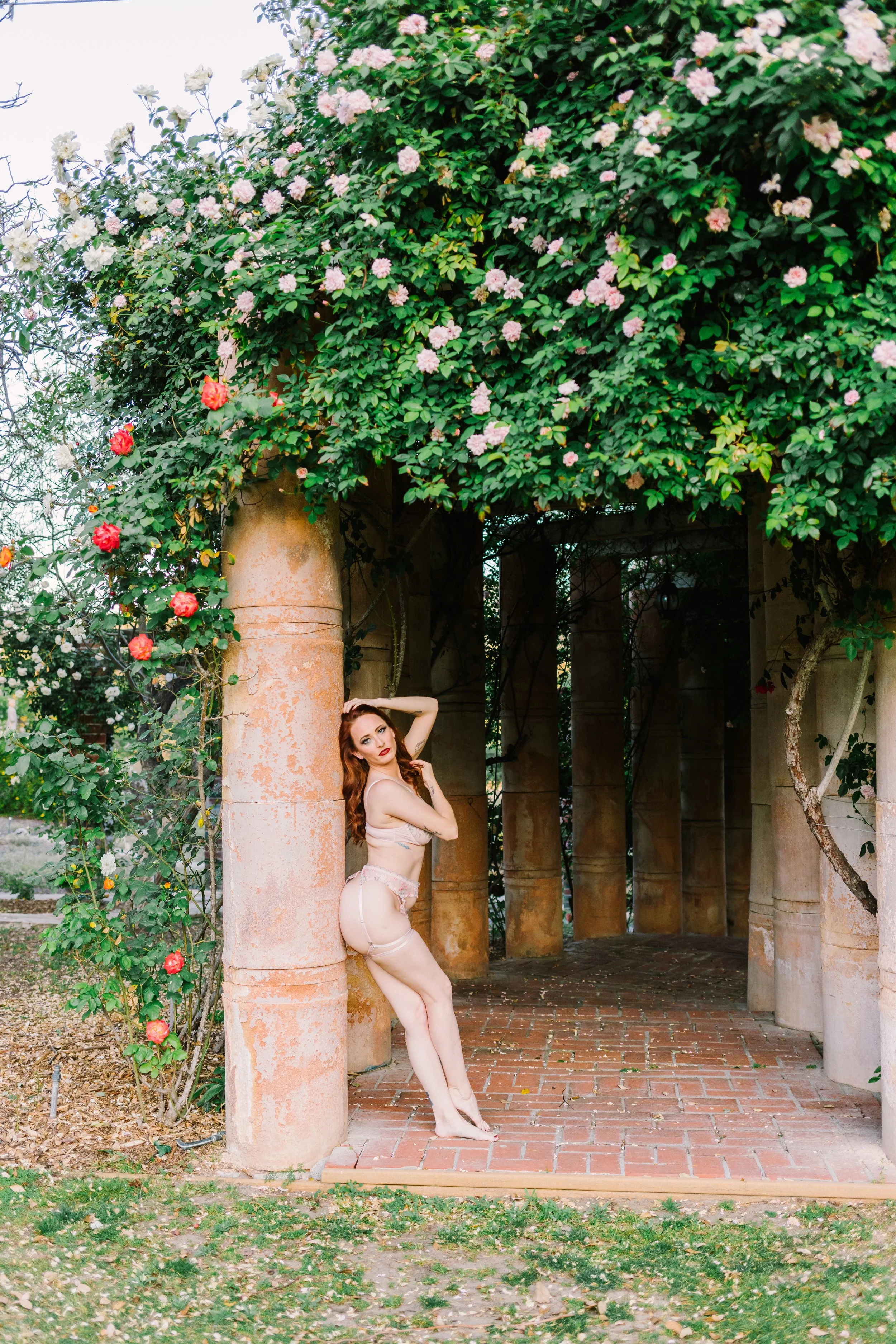 A woman with red hair and light-colored lingerie posing against a stone column in an outdoor garden with flowering bushes and a brick pathway.