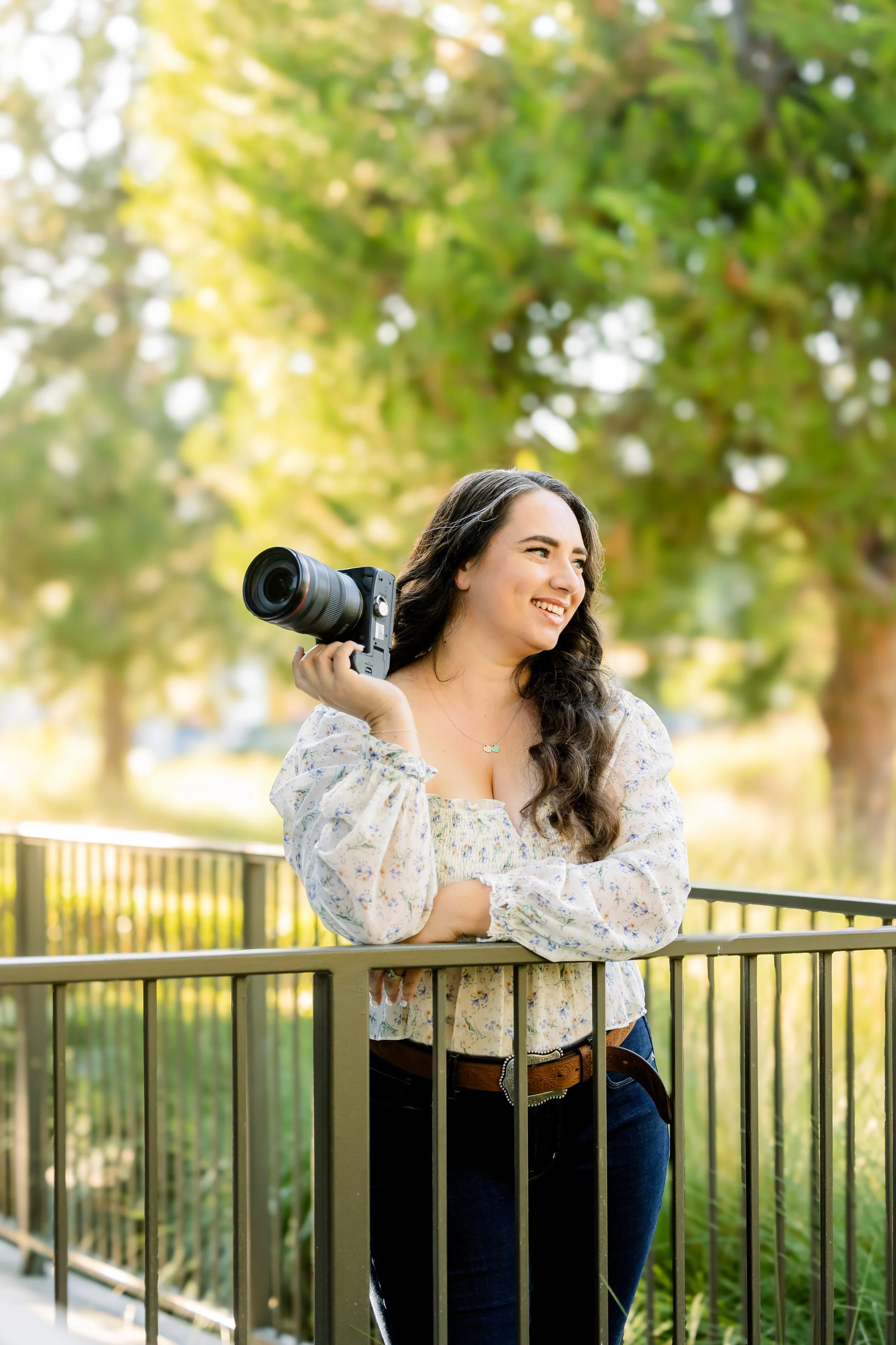 A woman with long dark curly hair, wearing a floral blouse and jeans, holding a camera on her shoulder, standing outdoors at a park with green trees and sunlight in the background.