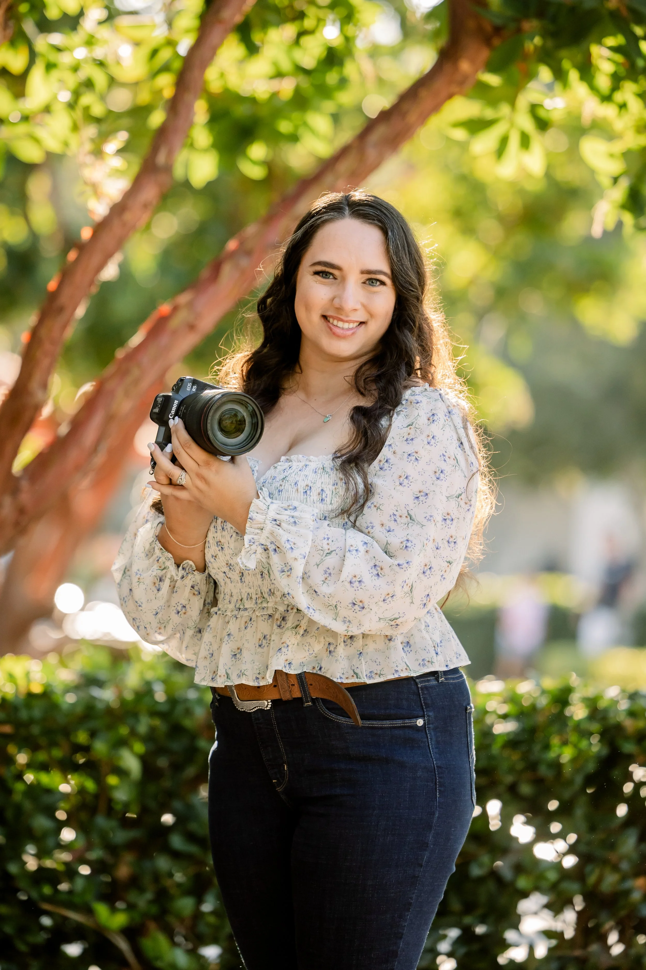 A young woman with long dark hair holding a professional camera outdoors in a garden with lush greenery and sunlight.