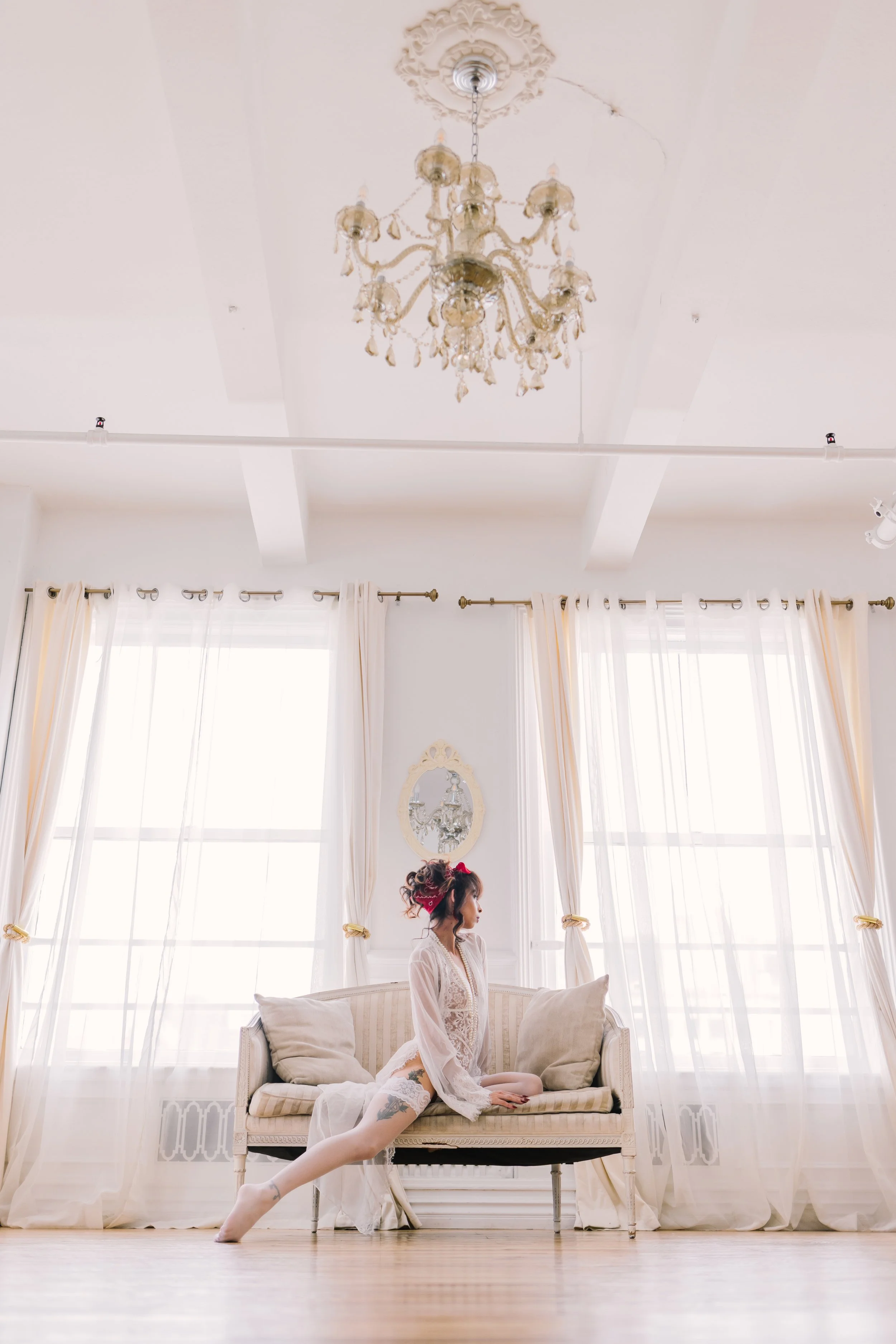 A woman with tattoos on her legs and curly hair in a messy updo with a red scrunchie, wearing a white lace robe, is sitting on a vintage cream-colored sofa with pillows in a bright room with large windows and sheer curtains, a chandelier hanging from the ceiling, and a mirror on the wall.