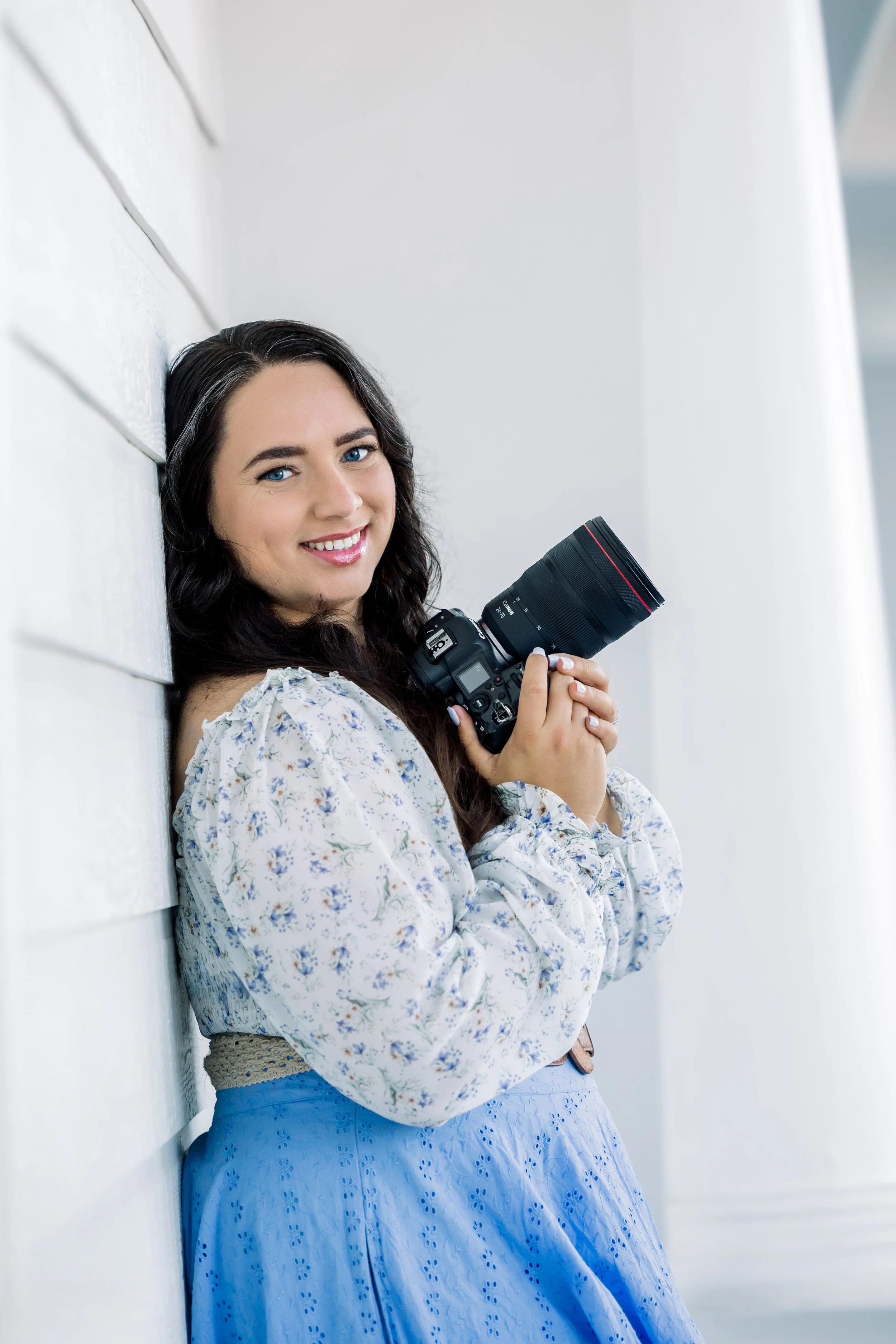 A woman with dark hair and blue eyes smiling, leaning against a white wall, holding a camera with a large lens.