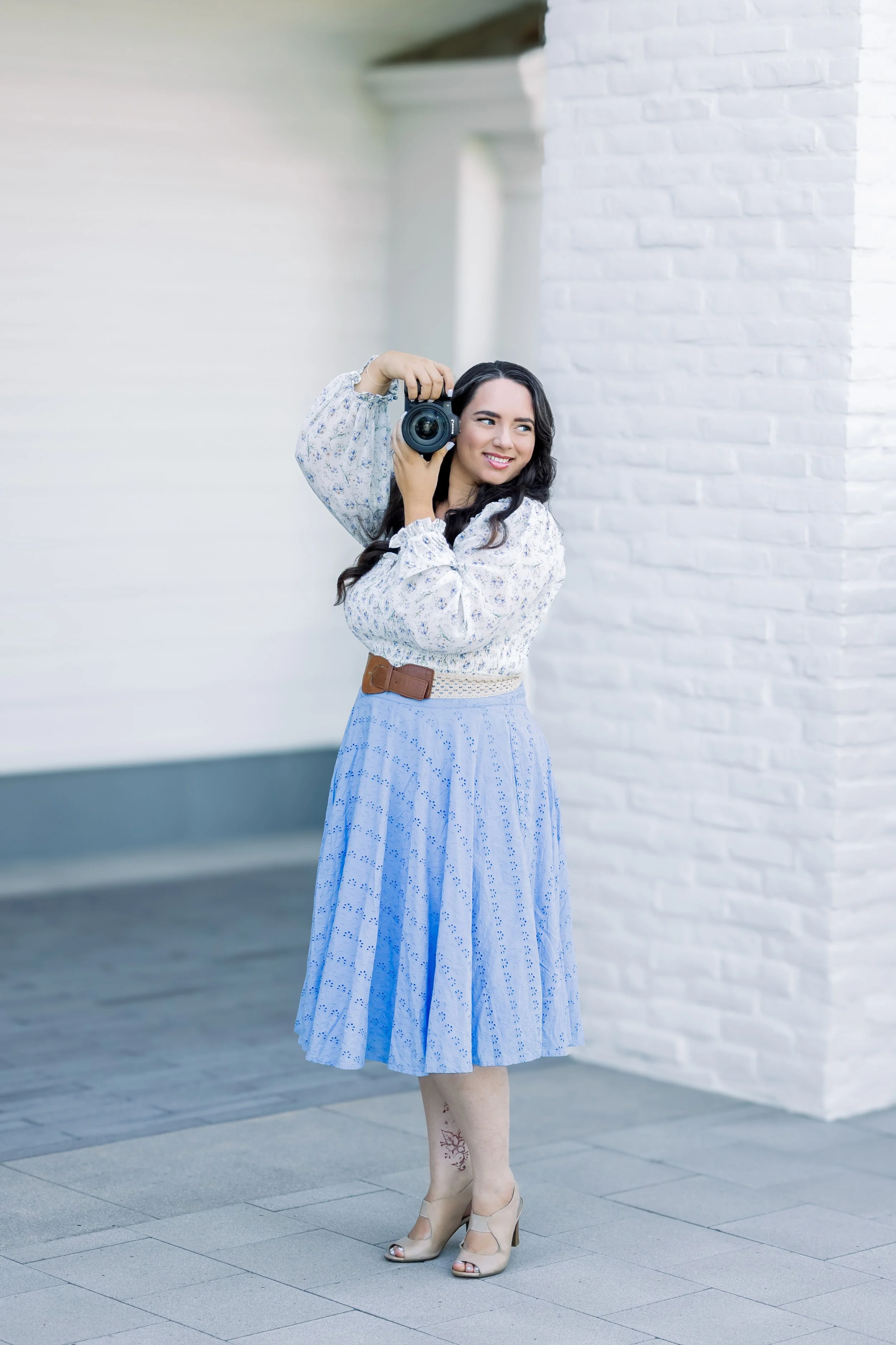A woman with long dark hair, wearing a white floral blouse, a blue pleated skirt, and beige high heels, is holding a camera to her shoulder while smiling against a white brick wall background.