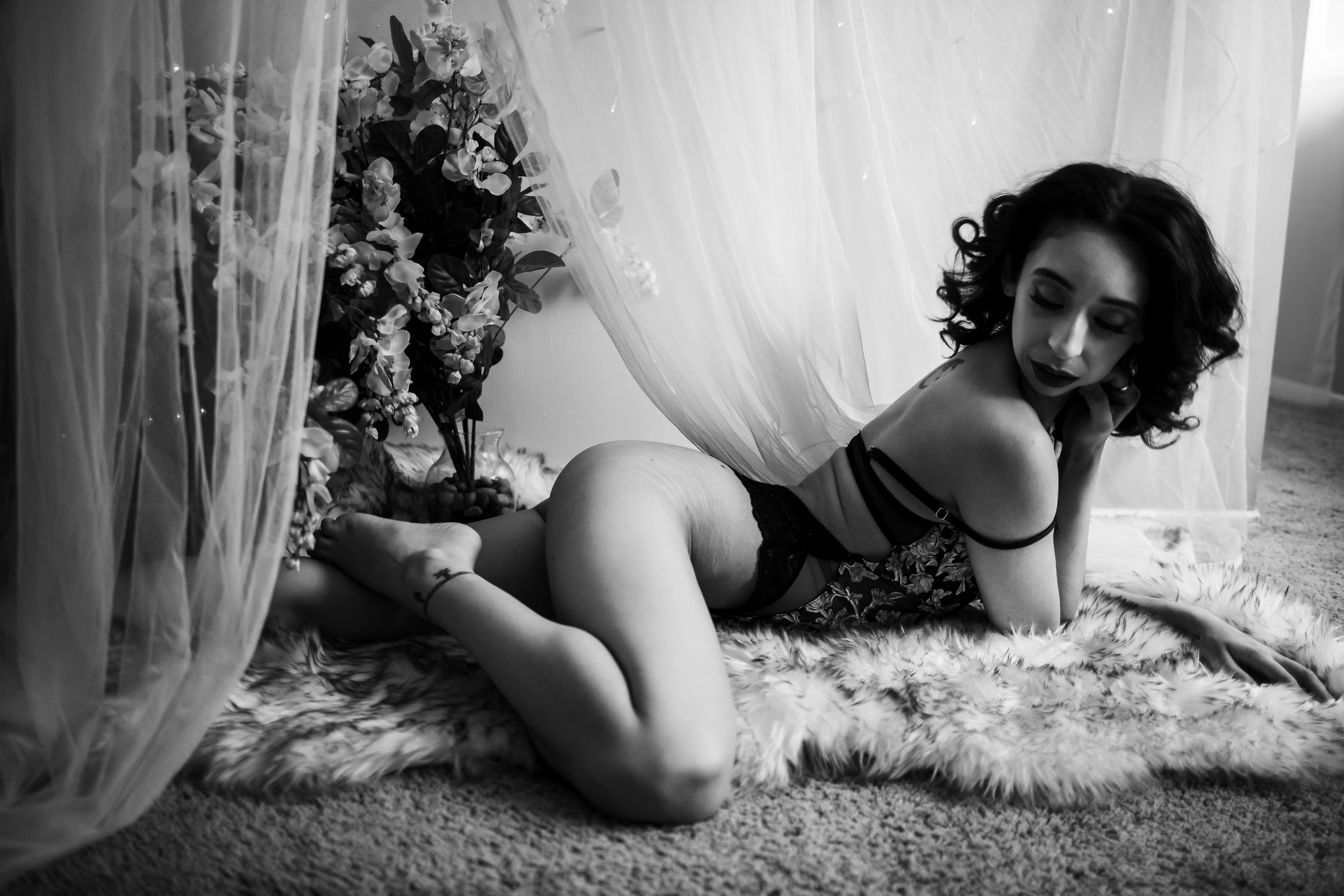 A woman with curly hair lying on her side on a fur rug, surrounded by sheer curtains and a floral arrangement, in black and white.