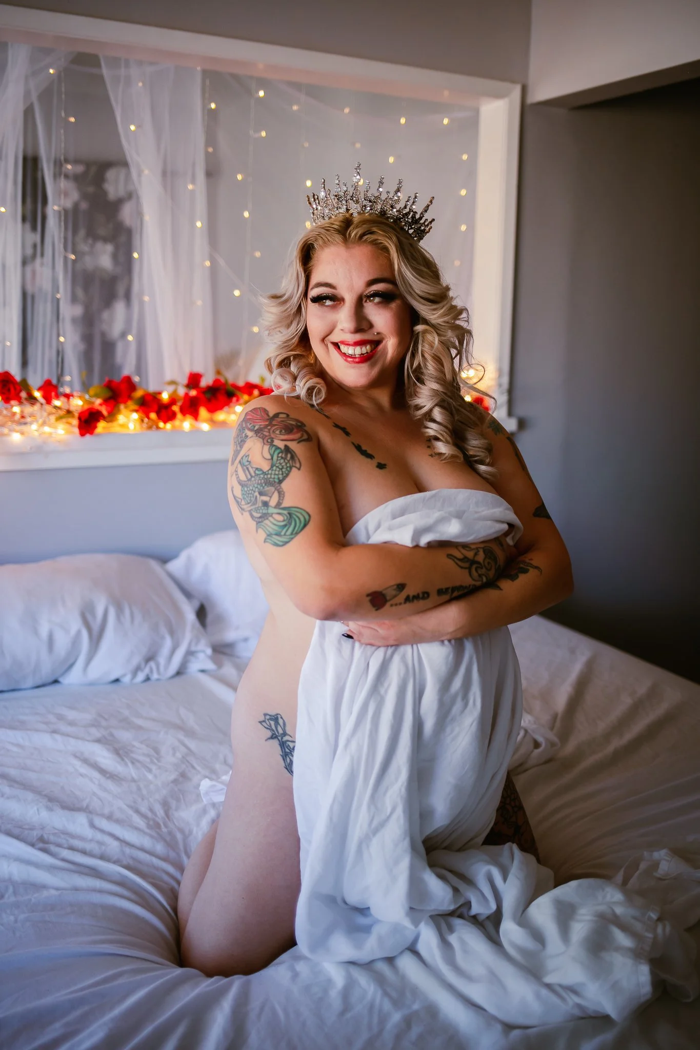 A woman with blonde hair and tattoos, wearing a tiara and a white sheet, sitting on a bed with white linens, smiling and looking to the side. The background includes fairy lights and pink flowers.