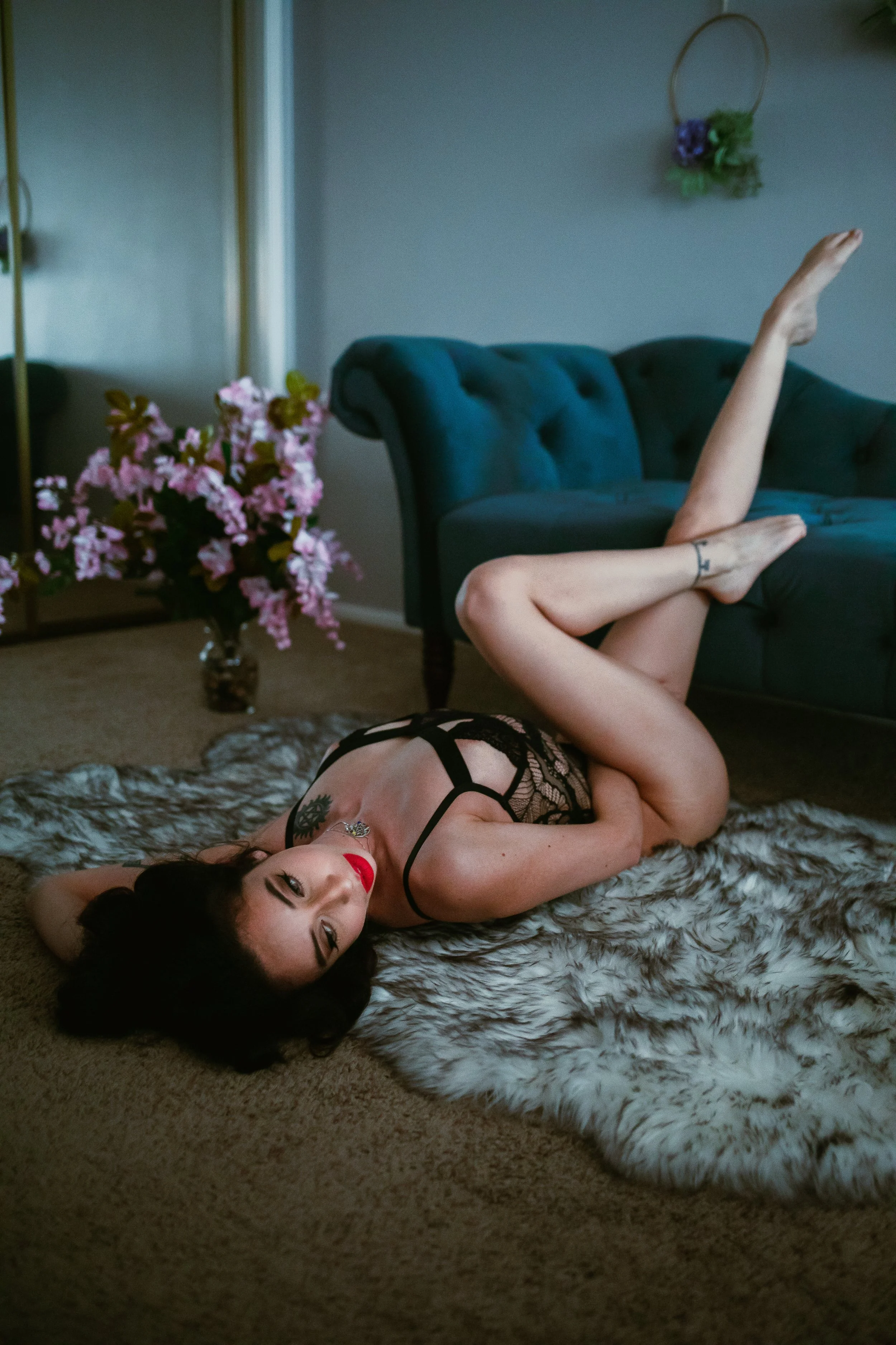 A woman lying on a faux fur rug on the floor with her head resting on her hand, wearing black lingerie, with one leg raised and resting on a teal couch in a living room decorated with pink flowers and wall hangings.