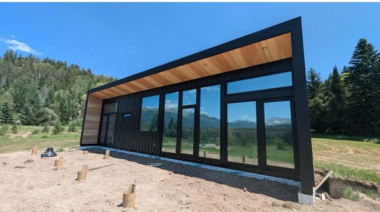 Modern house under construction with black exterior, large windows, and a wooden ceiling overhang, set against a backdrop of trees and mountains.