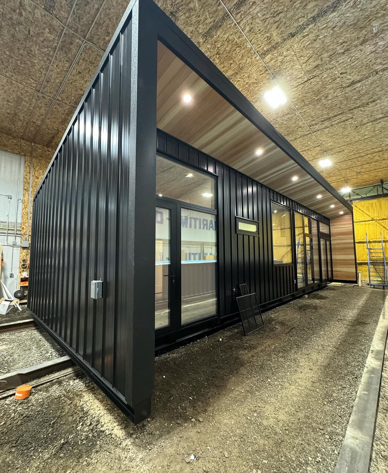 Modern shipping container-style building with black metal exterior, located indoors with ceiling lights, and an unfinished ground surface.