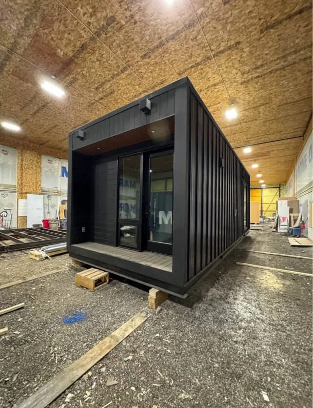 A small black living unit or office with a sliding glass door and a small front porch under construction inside a building with a wooden ceiling and unfinished walls.