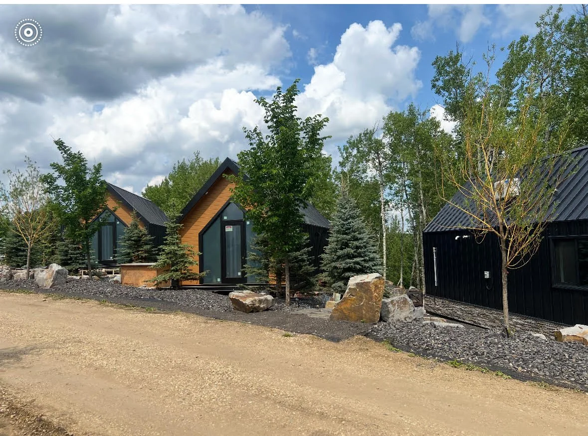 Three modern A-frame cabins with black and wood exteriors, surrounded by trees and rocks, under a partly cloudy sky.