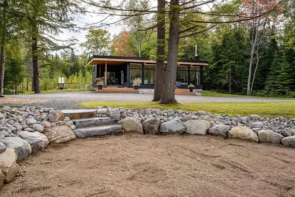 A modern house surrounded by trees with a yard that has rocks and a flat sandy area, possibly a garden or play area.