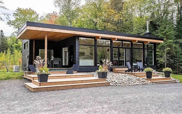 Modern black house with large glass windows, wooden decking with seating, surrounded by trees.