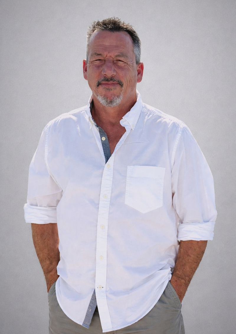 A middle-aged man with short, dark hair and a beard, wearing a white button-up shirt with rolled-up sleeves, standing against a plain light gray background.