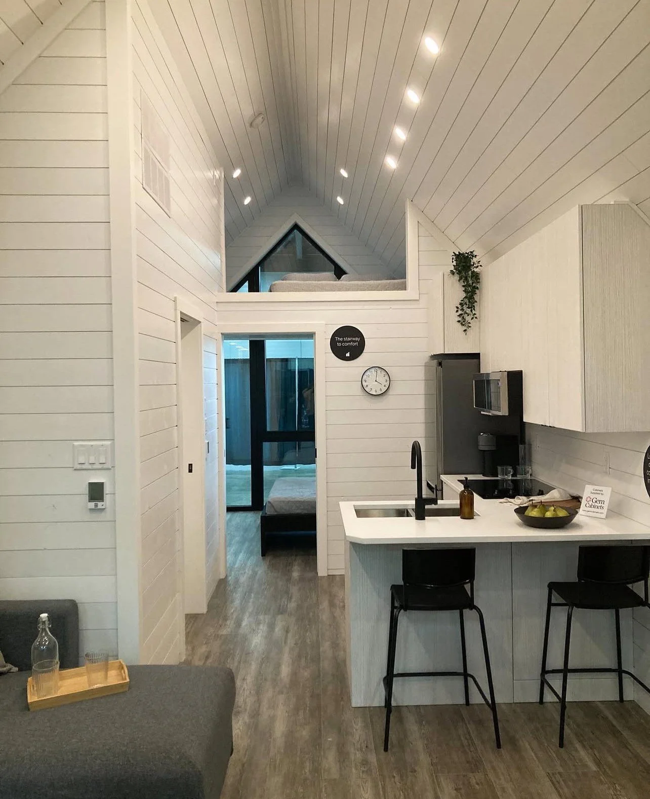 Interior of a modern attic apartment with white shiplap walls, a kitchen with black bar stools, a kitchen sink, and an upper sleeping loft with a bed, ceiling lights, and a triangular window.