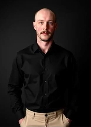 A man with a mustache and goatee, wearing a black button-up shirt and beige pants, standing against a dark background.