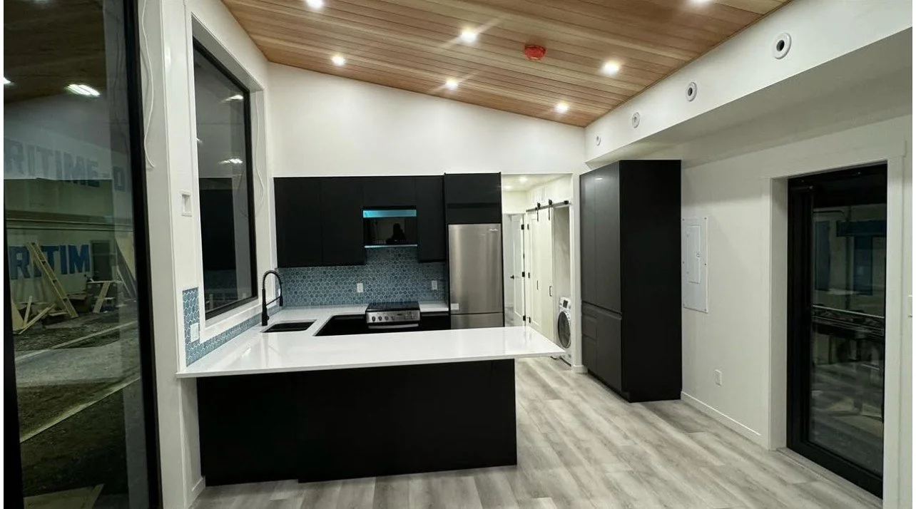 Modern kitchen with black cabinets, white countertops, stainless steel appliances, wood ceiling, and light-colored flooring.