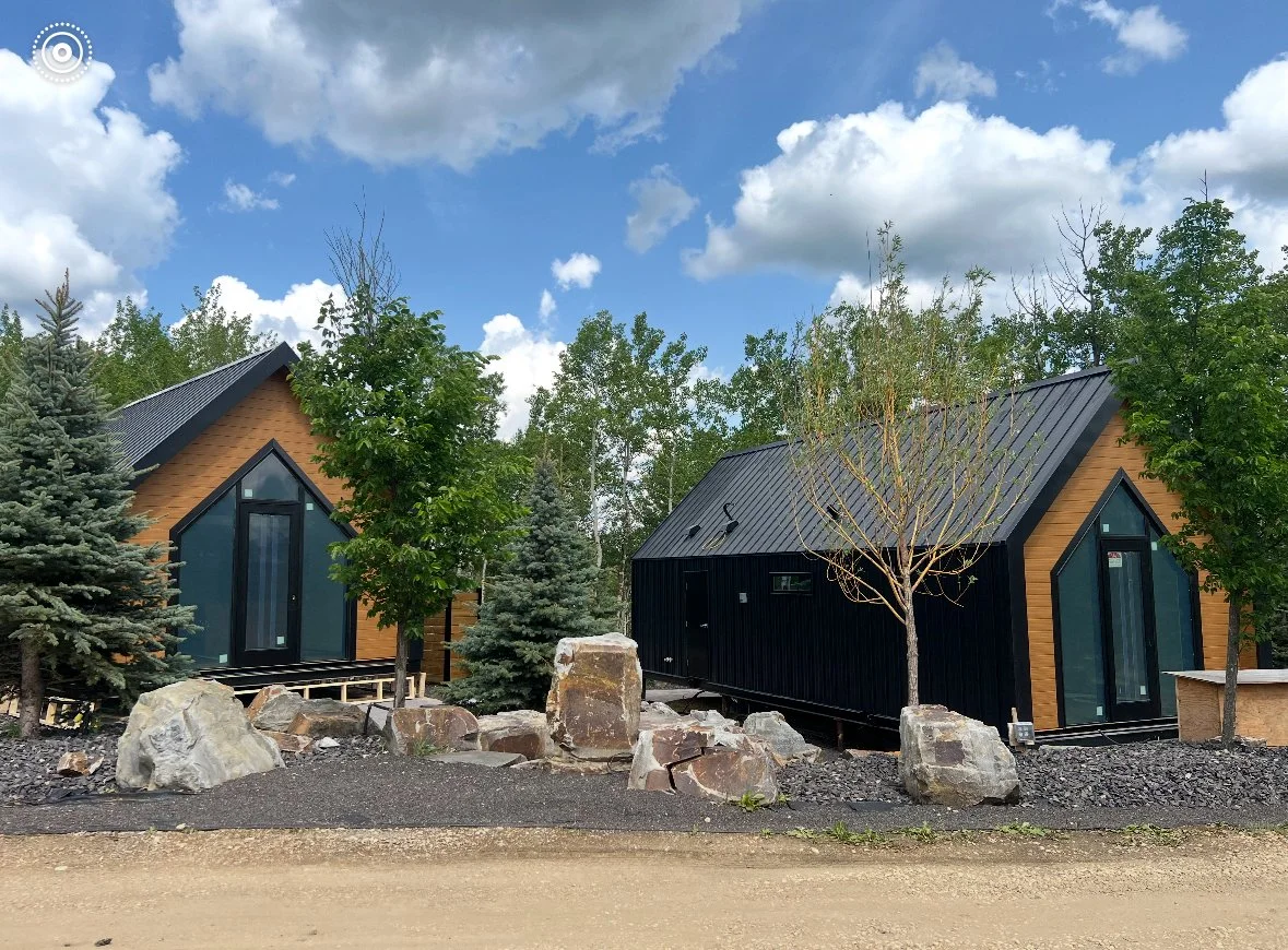 Two modern black and wood-paneled tiny houses with large glass doors, surrounded by trees, rocks, and a dirt road under a partly cloudy sky.