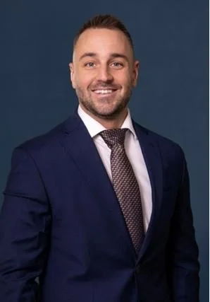 A man with a beard wearing a navy blue suit, white shirt, and patterned tie, smiling against a solid dark blue background.