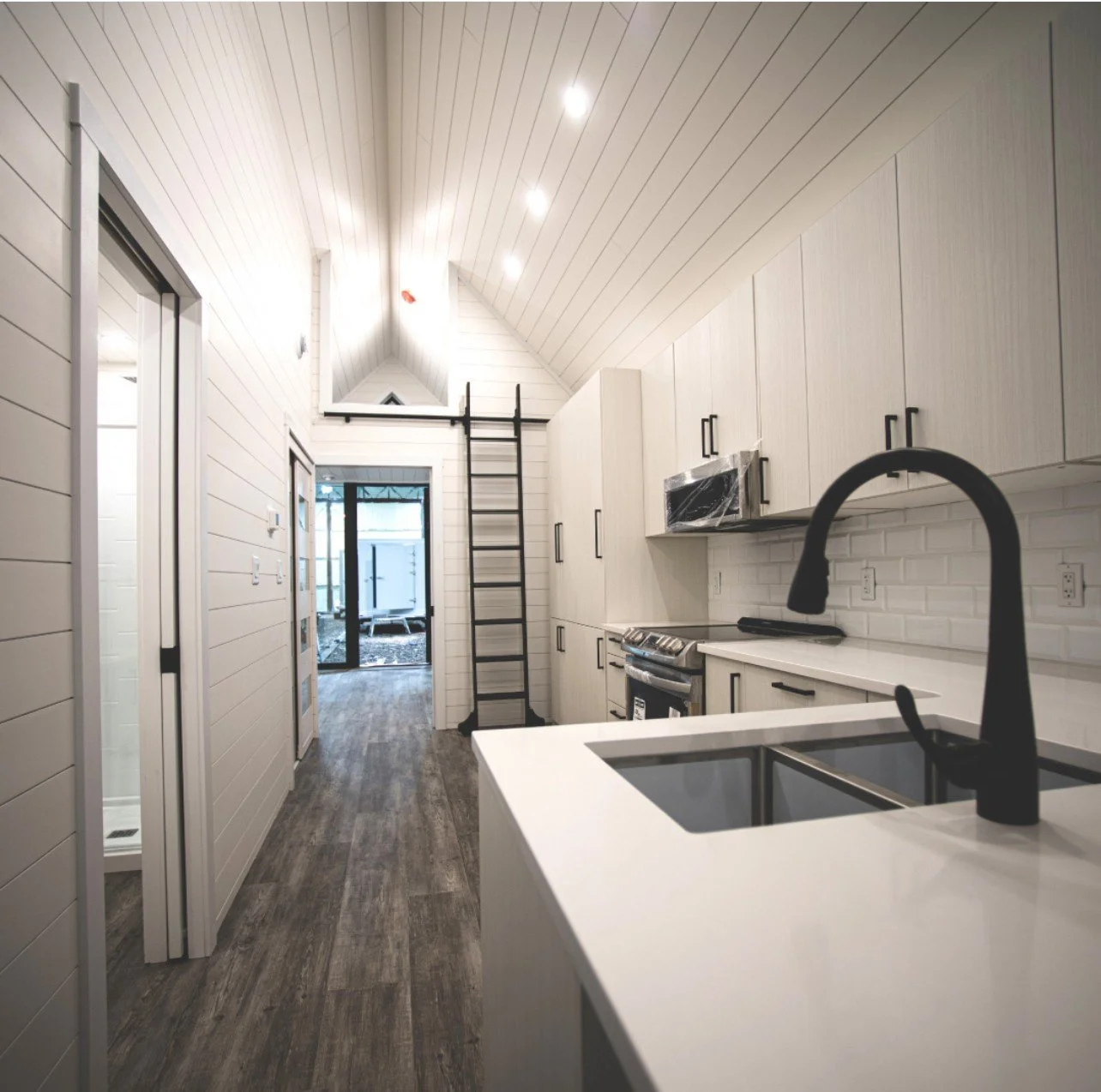 Modern kitchen with white cabinets, black hardware, gray wood flooring, and a black ladder leading to a small upper loft area