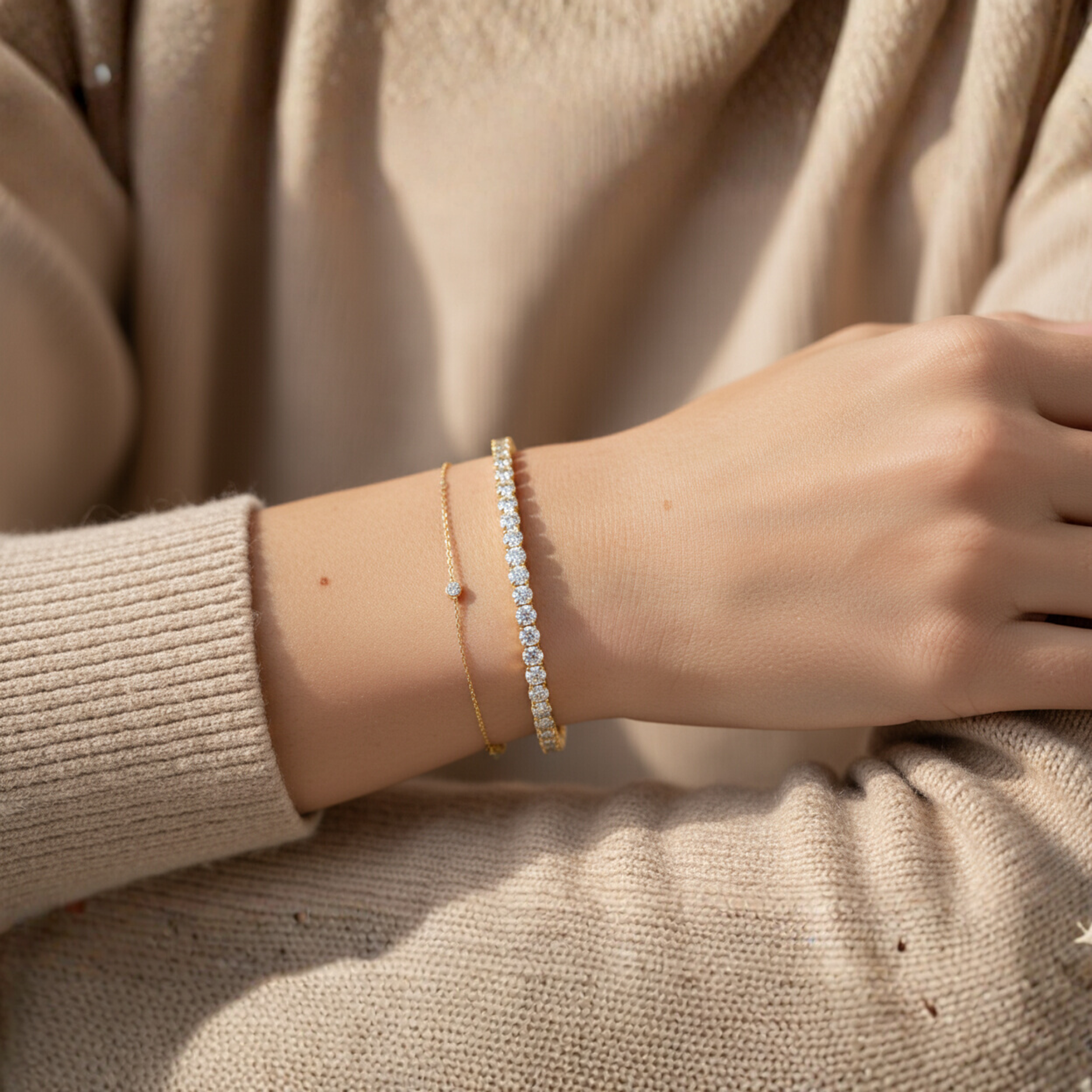 A person's arm resting on their lap, wearing beige clothing and two bracelets: a thin gold bracelet with small diamonds and a thicker diamond tennis bracelet.