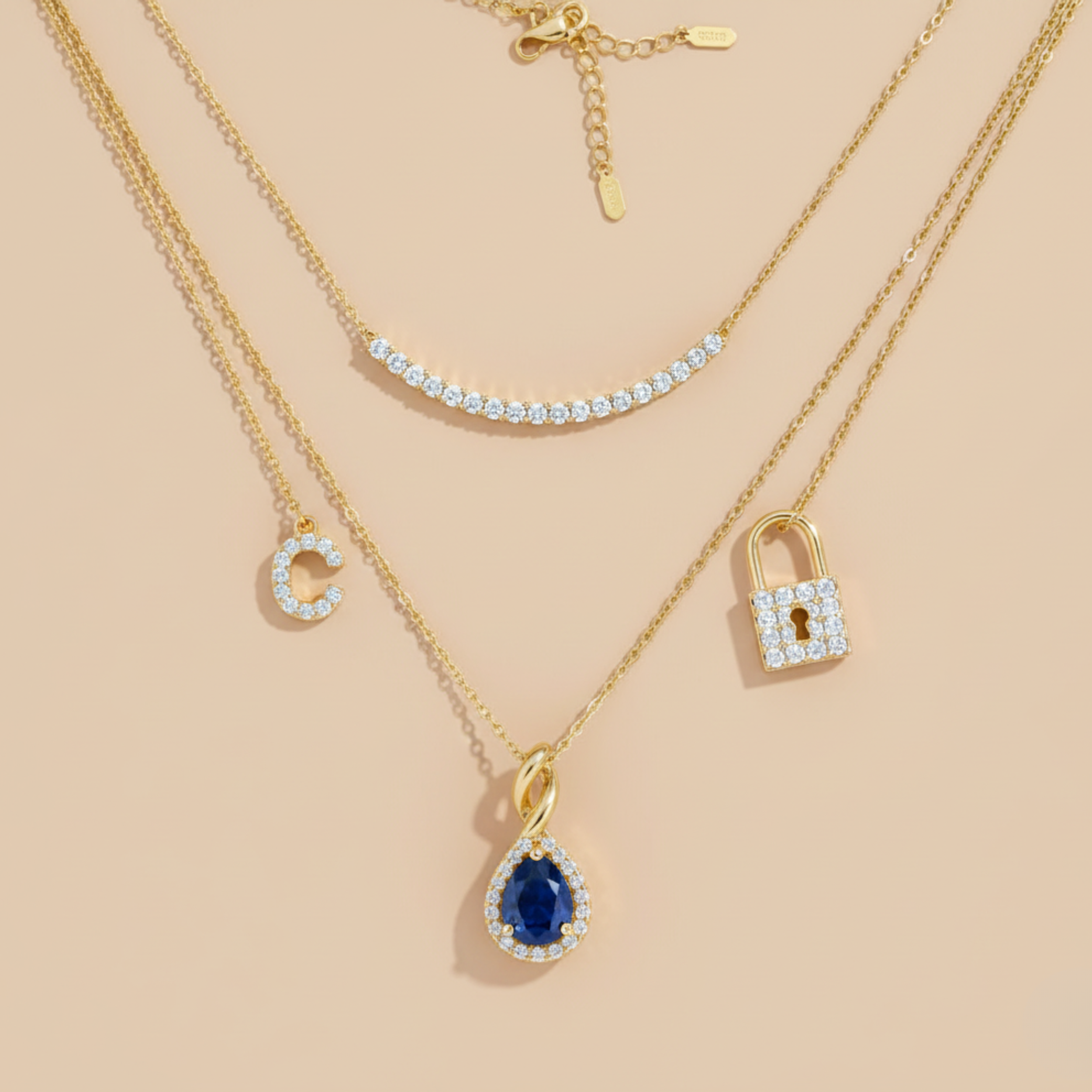 Three layered gold necklaces with diamond accents on a beige background. The top necklace has small diamonds, the middle has a pendant with a blue gemstone and diamonds, and the bottom has a diamond-encrusted lock charm and a diamond letter 'C' charm